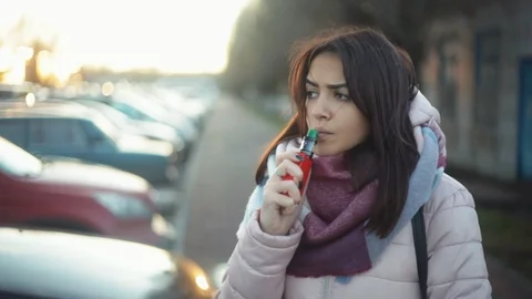 Pensive woman vaping e-cigarette between  cars on the parking zone near the Stock Footage 121483010