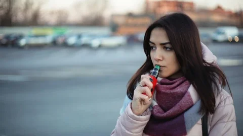 Pensive wooman vaping e-cigarette between  cars on the parking zone near the Stock Footage 121483098