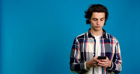 Pensive young man typing on smartphone and looking away isolated on blue Stock Footage 124535313