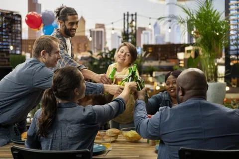 People clinking bottles of beer in group toast across rooftop dining table Stock Photos