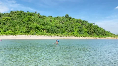 People crossing shallow river by walking along forest island beneath blue sky Stockbeeldmateriaal 345180703