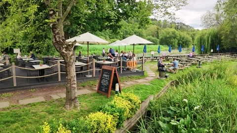 People enjoying drinks at a riverside pub with umbrellas and lush greenery Stock Footage 340107819