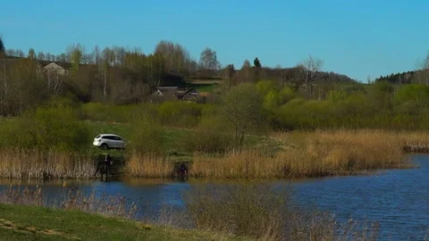 People fish by the water while a car is parked nearby on a sunny day in a rural Stock Footage 338425889