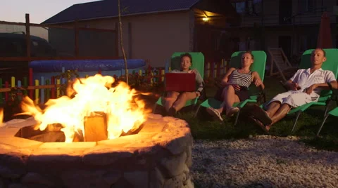 People relaxing in back yard sitting on patio loungers near fire pit at dusk Stock Footage 53046131