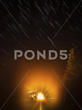 People seated around a campfire in the darkness, under a sky with star trails. Stock Photos