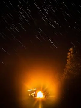 People seated around a campfire in the darkness, under a sky with star trails. Stock Photos