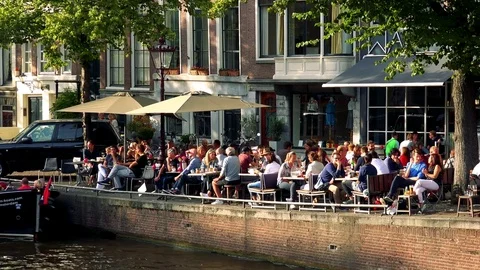 People sit at tables, some under parasols, on a street next to a river, Stock Footage 73958818