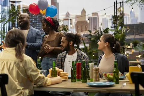 People socializing around rooftop dining table with food and bottled drinks Stock Photos