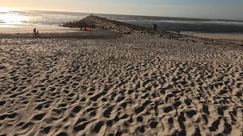 People walk along a sandy beach toward a rocky breakwater Stock Footage 346290554