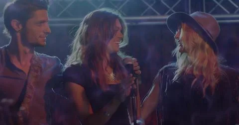 Performing trio exchanging smiles on dim stage, with microphone, hat, spotlights Stock Photos