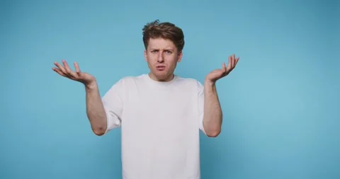 Perplexed young man stands against blue backdrop with confused expression Stock Footage 313280562