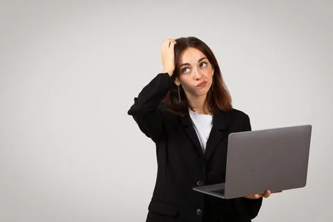 Perplexed young professional woman holding a laptop, scratching her head with a Stock Photos