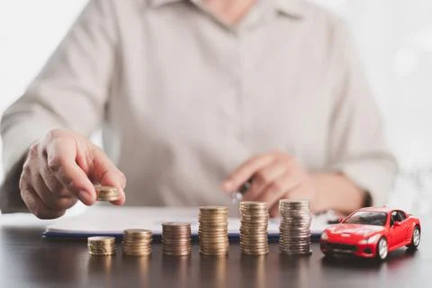 Person adding coins to rising stacks beside a red model car, representing v.. Stock Photos