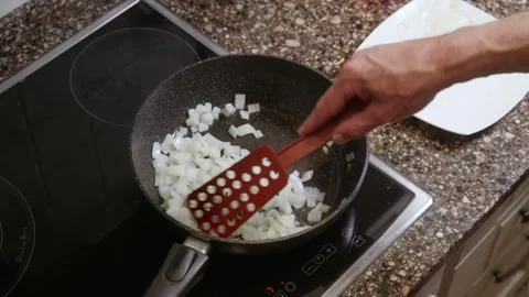 Person adding diced onion to black frying pan on stove with red spatula Stock Footage 323901842