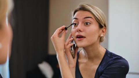 A person applies eye makeup while engaged in a phone conversation in a cozy Stock Footage 337555322