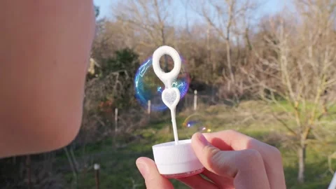 A person blowing a large iridescent soap bubble with a bubble wand outdoors. Видео 330362017