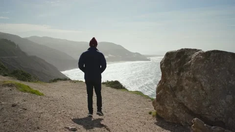 Person in blue jacket walks to the edge of a cliff overlooking a coastal ocean. Stock Footage 150137181