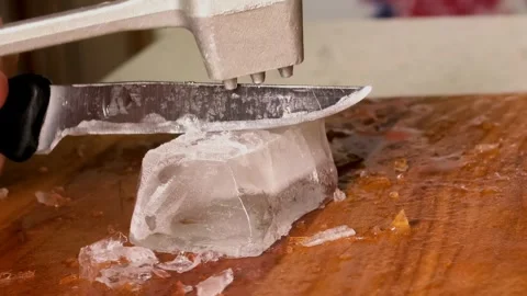Person breaks a large ice cube with a knife and kitchen mallet on a wooden Stock Footage 342718476