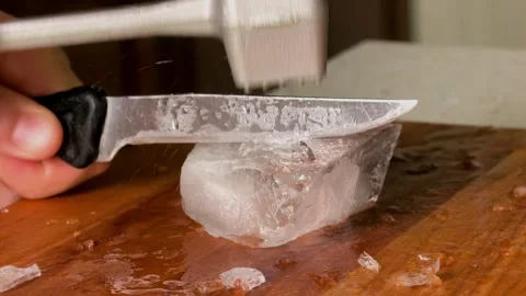 Person breaks a large melting ice block with a kitchen knife and hammer on a Stock Footage 340189857