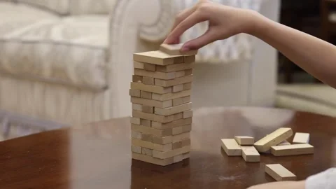 A person carefully pulls a block from a tall stack of wooden blocks on a table Stock Footage 278887817