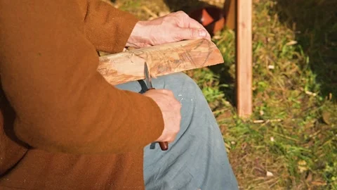 A person is carving wood using a simple handheld tool while seated outdoors. The 库存影片 345033048