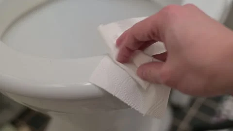 A person cleans the rim and surface of a toilet bowl with a paper towel. The 库存影片 337101340