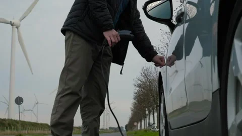 Person connecting charging cable to electric car beside rural road with wind Stock Footage 341042399