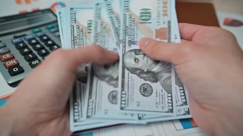 A person counts cash while seated at a desk. The workspace has financial charts, 库存影片 338301487