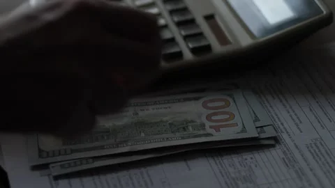 A person counts a stack of hundred-dollar bills using a calculator next to .. Stock Footage 348276305
