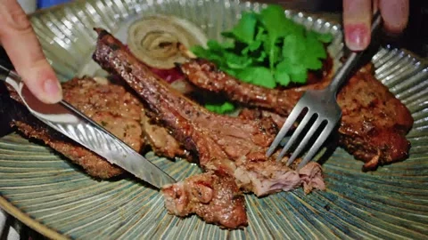 Person cutting grilled lamb chops with fork and knife on ceramic plate in r.. Vídeos de archivo 339754742