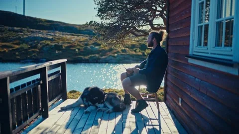 A Person Eats a Snack on a Cabin Deck While a Dog Rests Nearby by the Lake - Stock Footage 336122248