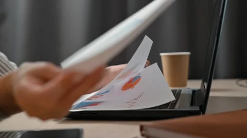 A person engages in work by reviewing documents and typing notes on a laptop in Stock Footage 339005554