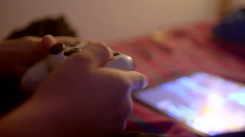 A person is engaging with a game controller while using a tablet, with a bed in  Stock Footage 295932984