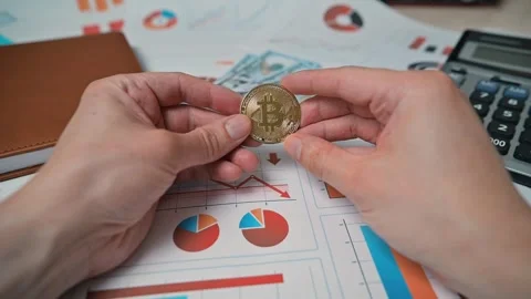 A person examines a bitcoin token while sitting at a desk filled with charts and Stock Footage 338301513