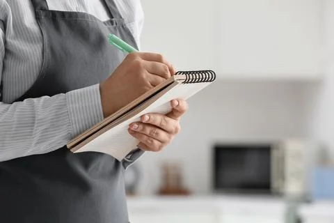 Person in a gray apron writing notes in a spiral-bound notebook with a green  Stock Photos