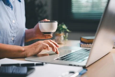 Person holding a coffee cup while using a laptop at a home workspace, repre.. Stock Photos
