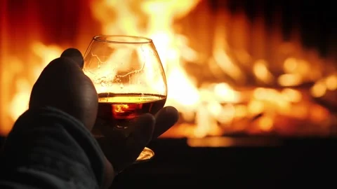 Person holding glass of brandy in front of warm fireplace on cozy winter ev.. Stock Footage 334594053