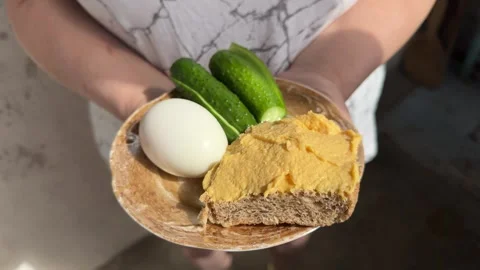Person holding plate with boiled egg, fresh cucumbers and brown bread  Stockbeeldmateriaal 345327626