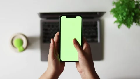 Person holding a smartphone with a green screen in front of a laptop Stock Footage 337173353