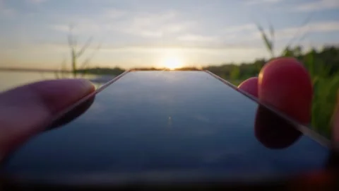 Person Holding Smartphone Toward Setting Sun Along Lake at Sunset Stock Footage 345417582