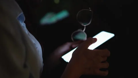 A person is holding a smartphone while in dim lighting, with glasses held in the Stock Footage 289965119