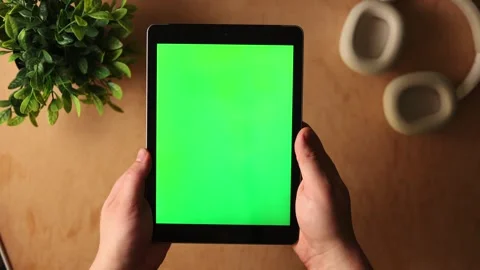 Person holding a tablet with a green screen on a wooden desk with headphones and Stockbeeldmateriaal 341737575