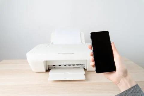 Person holds a smartphone in front of a printer on a wooden desk, wireless mo Stock Photos