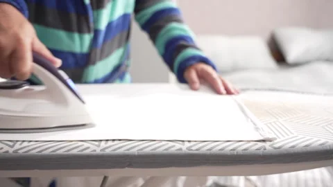 Person irons a white shirt on a patterned board, holding fabric steady. 스톡 동영상 314957137