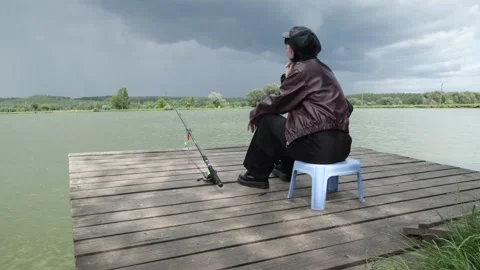 A person in a leather jacket and cap sits alone on a pier, fishing calmly while Stock Footage 311803523