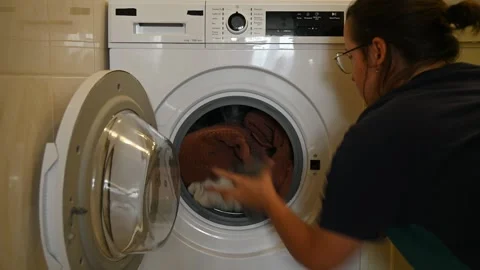Person loading laundry into front load washing machine in laundry room Stock Footage 339664496