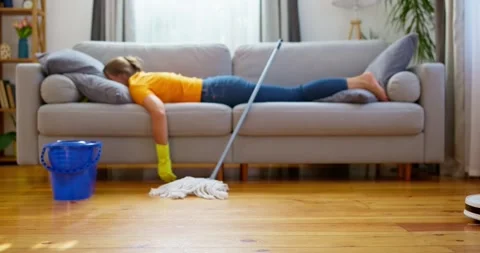 Person lying on sofa with mop, robotic vacuum cleaner on hardwood floor. Stock Footage 269764276