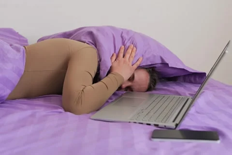 A person is lying on their side in bed, using a laptop. They cover their face Stock Footage 336464086