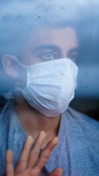 Person with a mask on his face while looking out the window with water drops. Stock Photos