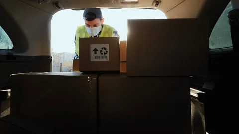 A person in a mask puts boxes in the trunk of a car. Volunteer work in the midst Stock Footage 129293545